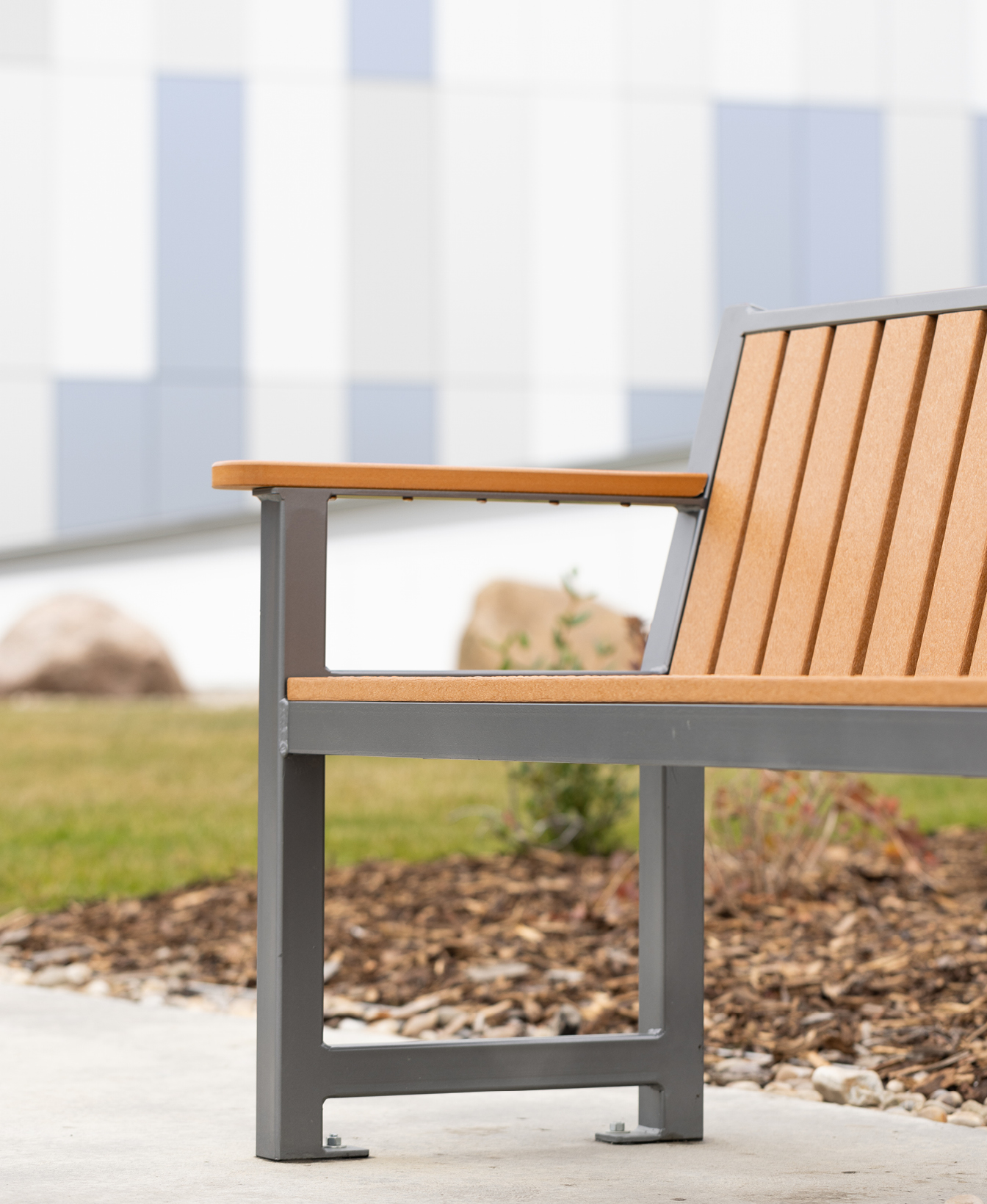 A close-up of the 400 Backed Bench, featuring wooden slats and a metal frame, placed on concrete near landscaped mulch and grass.