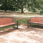 Two wooden benches with metal frames sit on a wooden deck in a shaded park area with trees and grass in the background.
