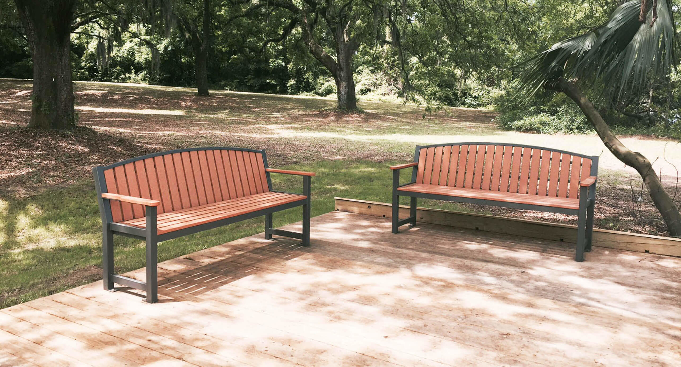 maglin_mbe-0450-backed-benches_1 Two wooden benches with metal frames sit on a wooden deck in a shaded park area with trees and grass in the background.