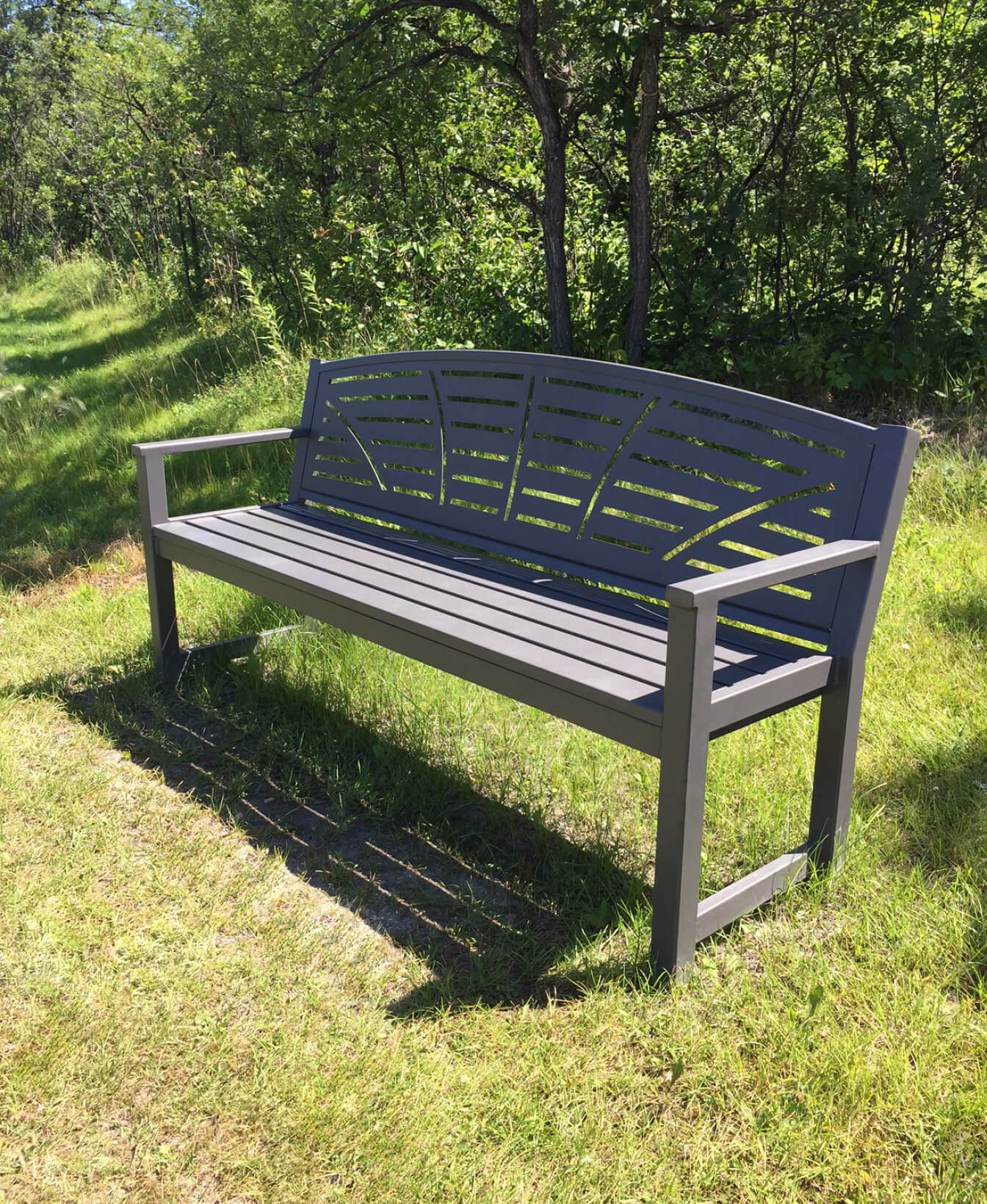 The 450 Backed Bench, featuring a gray metal frame and slatted backrest, stands on grass amidst lush green trees and sunlight.