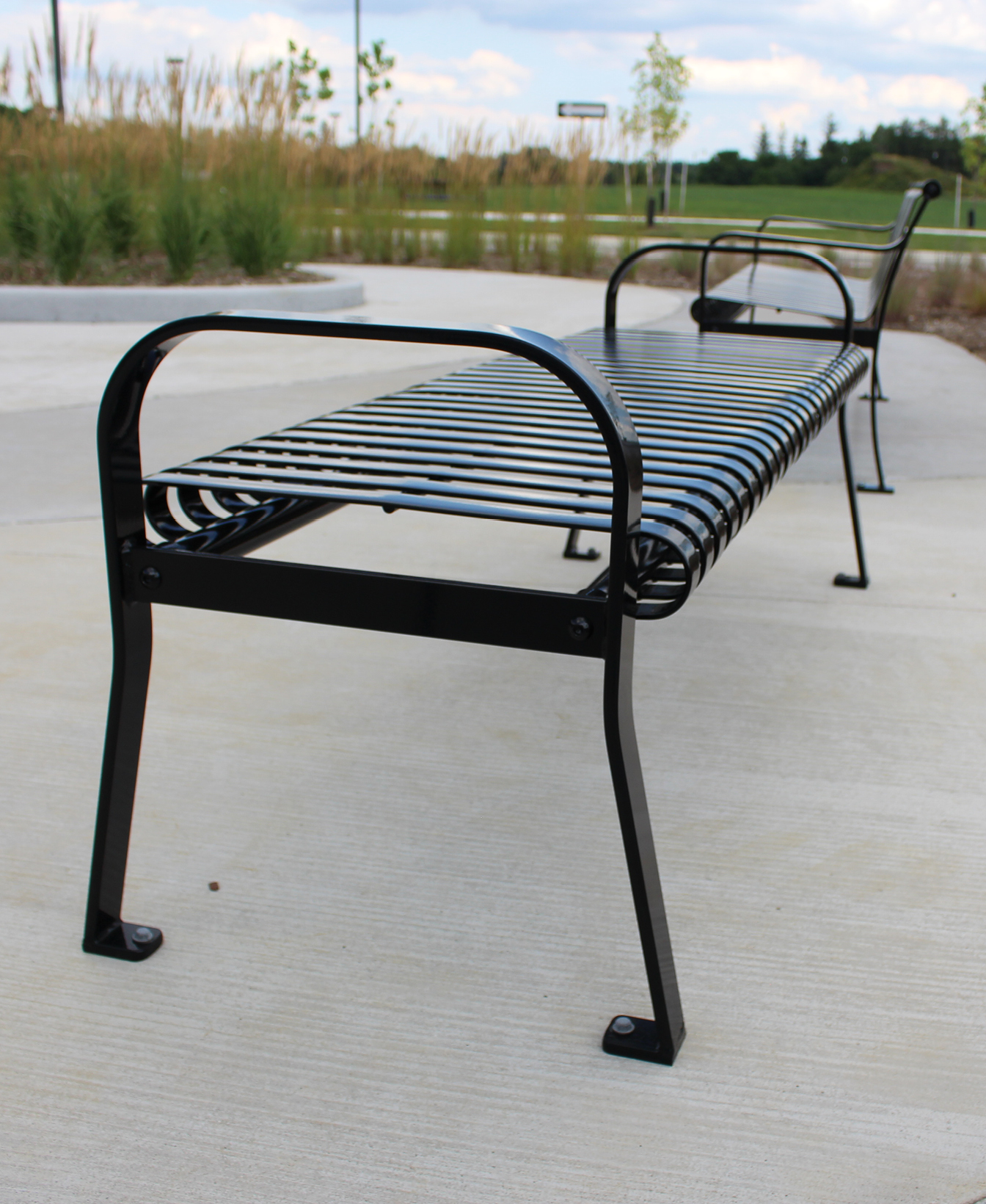 The 510 Backless Bench, made of black metal with a slatted seat, is set on a concrete surface in a landscaped park area.