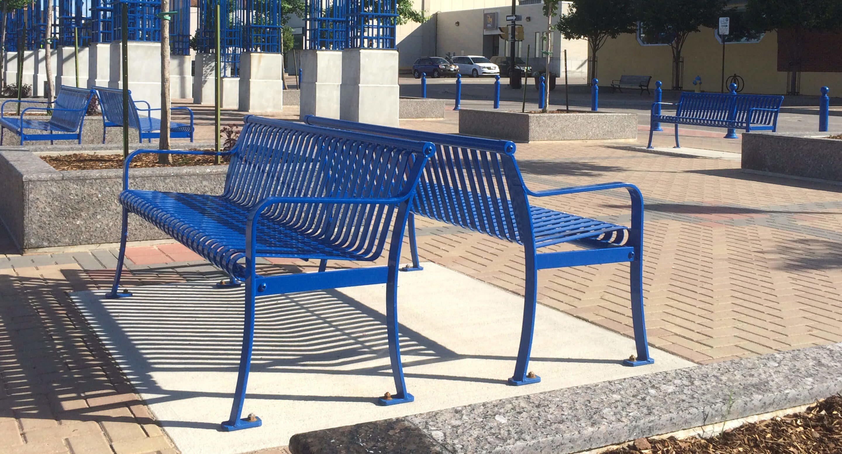 maglin_mbe-0510-backed-benches_1 A blue double-sided metal bench is positioned on a brick walkway in an urban outdoor setting, with other benches and street features visible in the background.