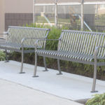 Two metal benches are placed on a concrete sidewalk next to a building with glass windows and surrounded by green plants.