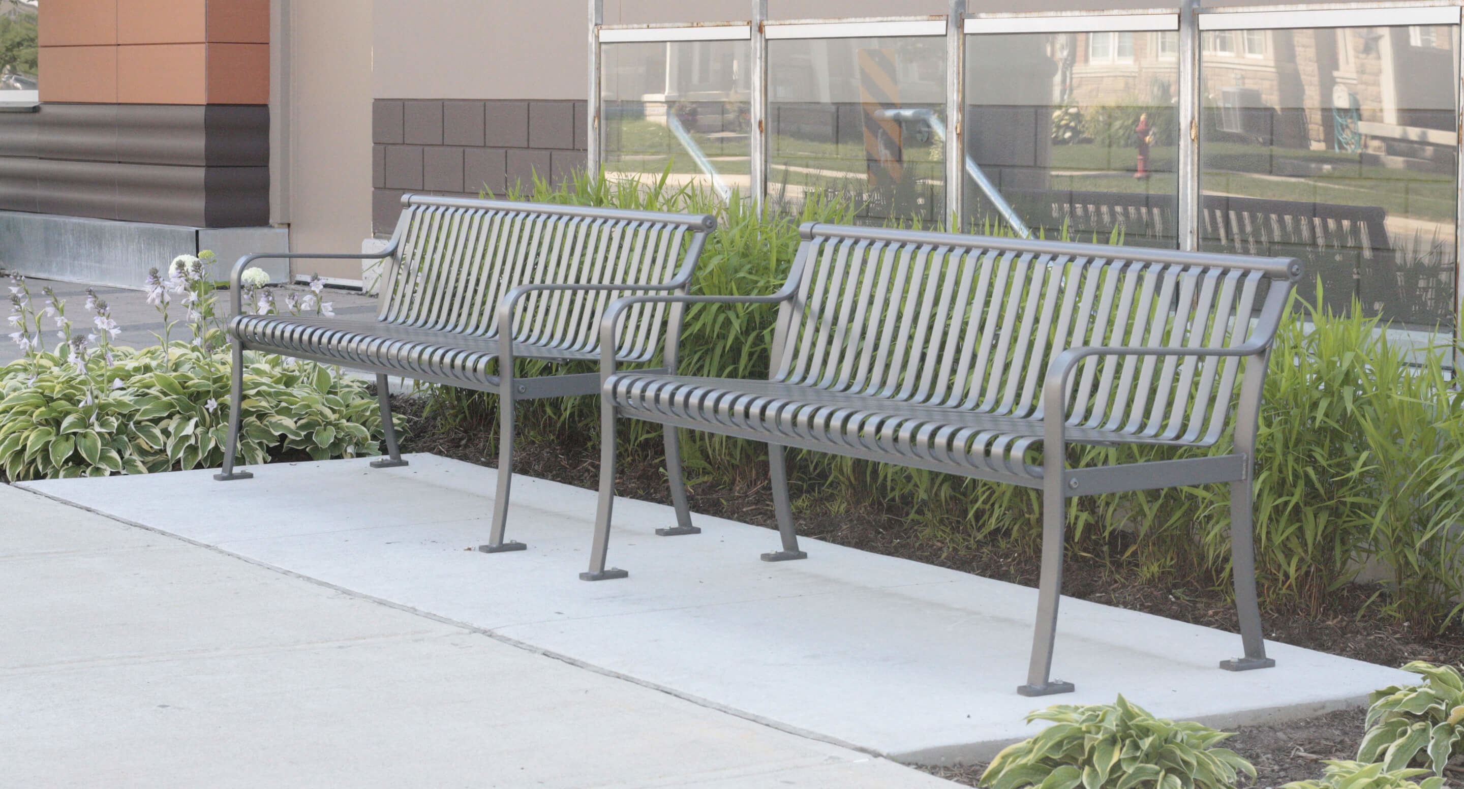 maglin_mbe-0510-backed-benches_2 Two metal benches are placed on a concrete sidewalk next to a building with glass windows and surrounded by green plants.