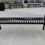 Black metal slatted bench with no backrest on a concrete surface in an outdoor urban area, with other benches and a trash can in the background.