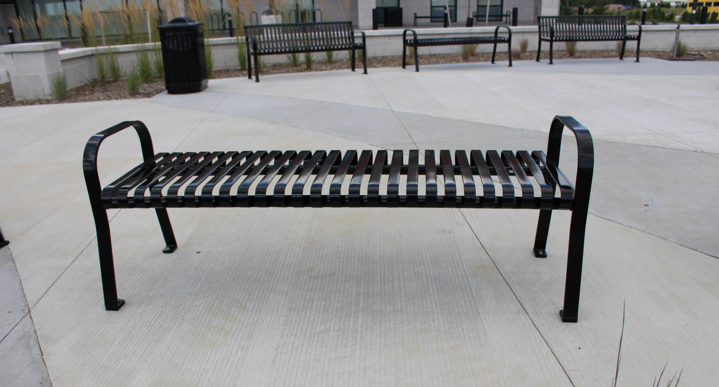maglin_mbe-0510-backless-benches_1 Black metal slatted bench with no backrest on a concrete surface in an outdoor urban area, with other benches and a trash can in the background.