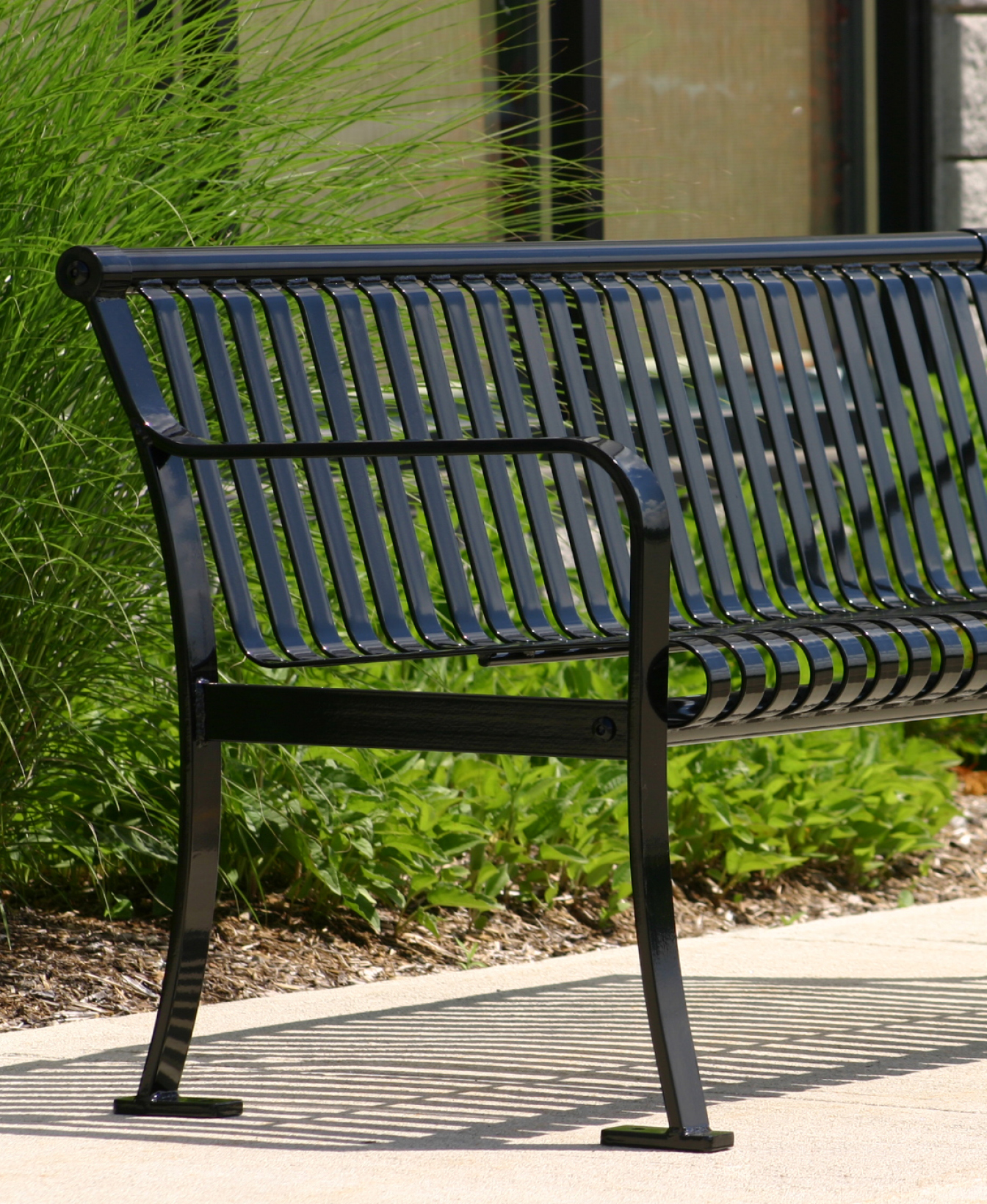 A 510 Backed Bench in black metal sits on a concrete surface beside green plants and tall grass.