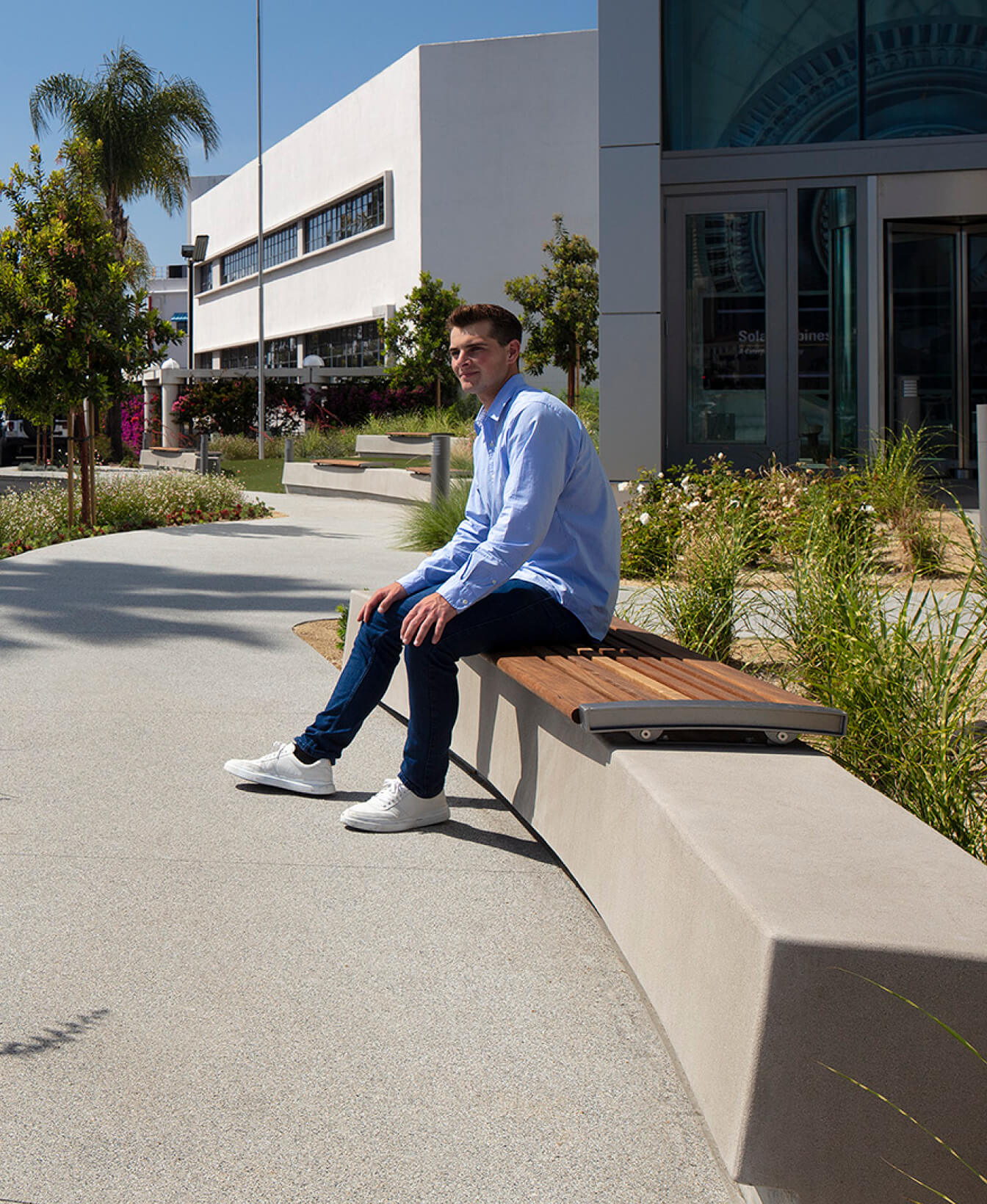 A man in a light blue shirt and jeans sits on a 720 Backless Bench near a building with glass doors and landscaped greenery.