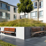 A modern courtyard features a raised grassy mound with built-in wooden benches and is surrounded by apartment buildings and paved walkways.