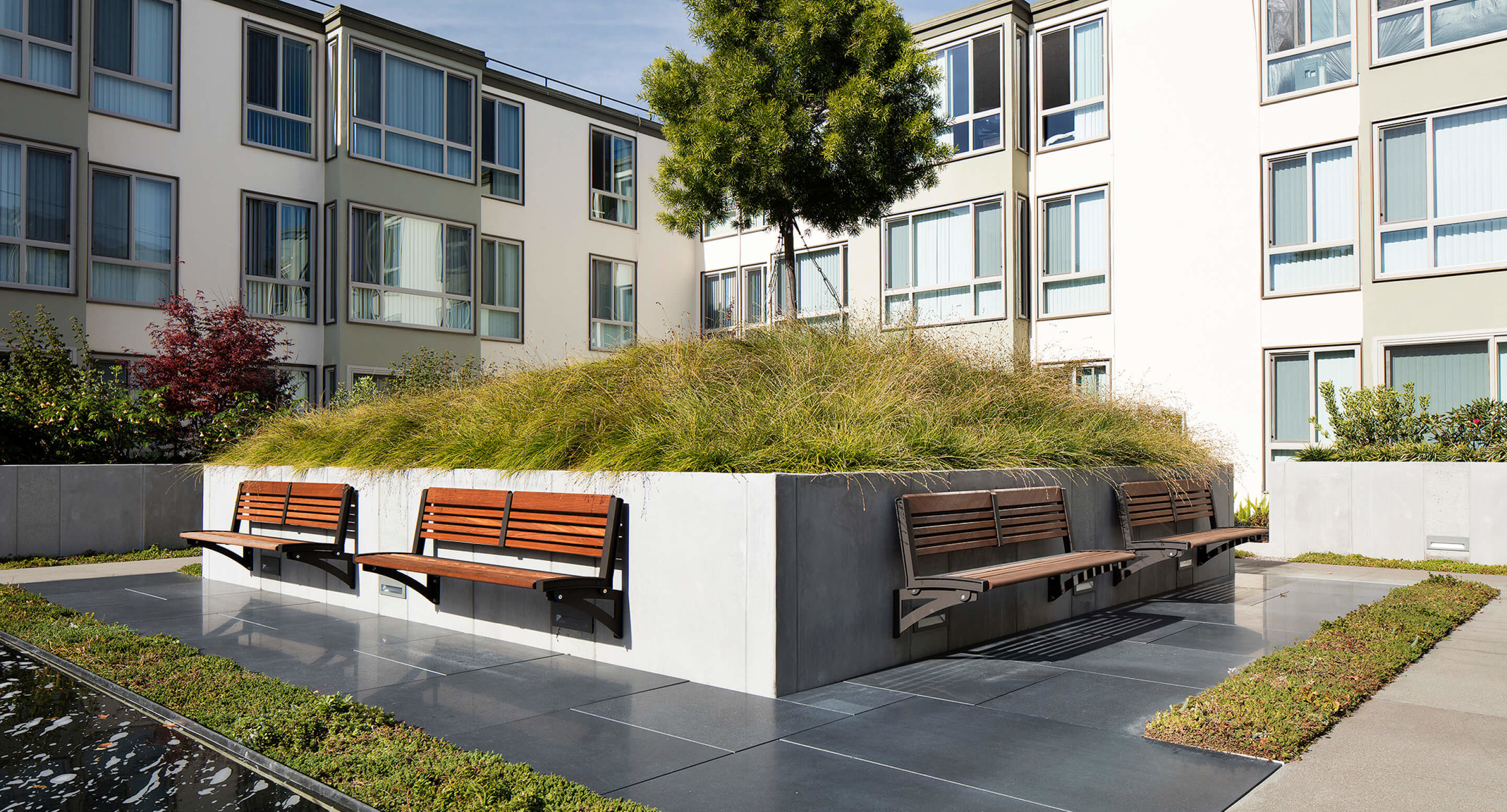 A modern courtyard features a raised grassy mound with built-in wooden benches and is surrounded by apartment buildings and paved walkways.