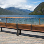 Two wooden benches on a paved area overlook a lake with mountains and trees in the background under a partly cloudy sky.