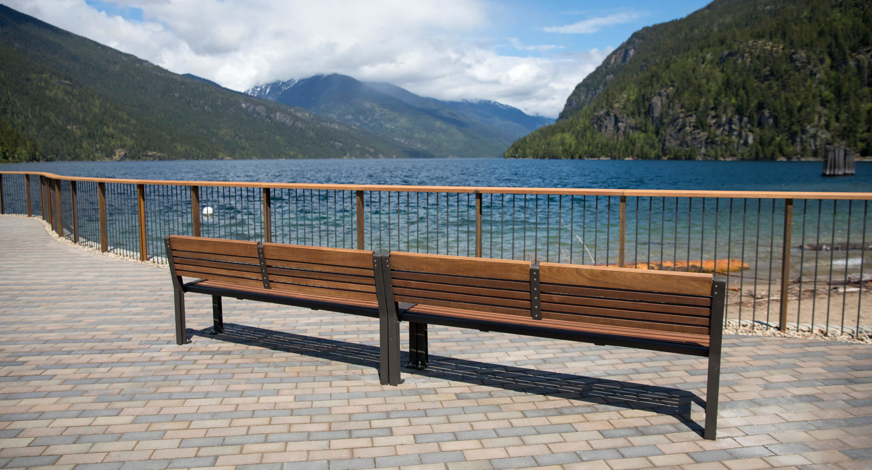 Two wooden benches on a paved area overlook a lake with mountains and trees in the background under a partly cloudy sky.