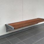 A wooden bench with metal brackets is mounted on a white wall above a gray tiled floor.