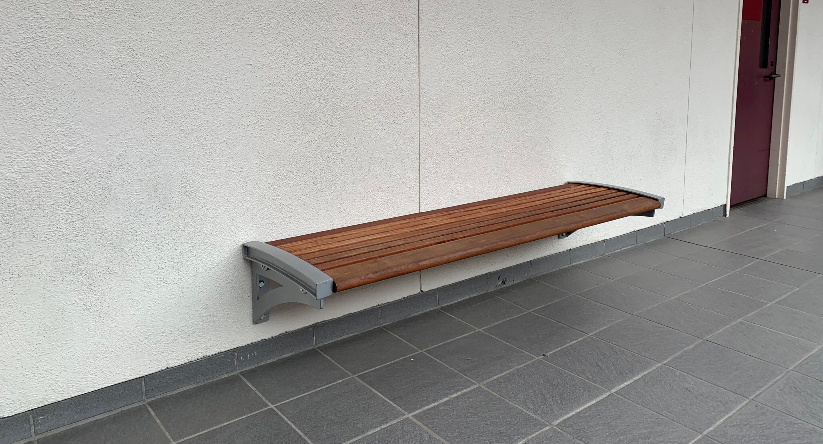 maglin_mbe-0720-backless-benches_3 A wooden bench with metal brackets is mounted on a white wall above a gray tiled floor.
