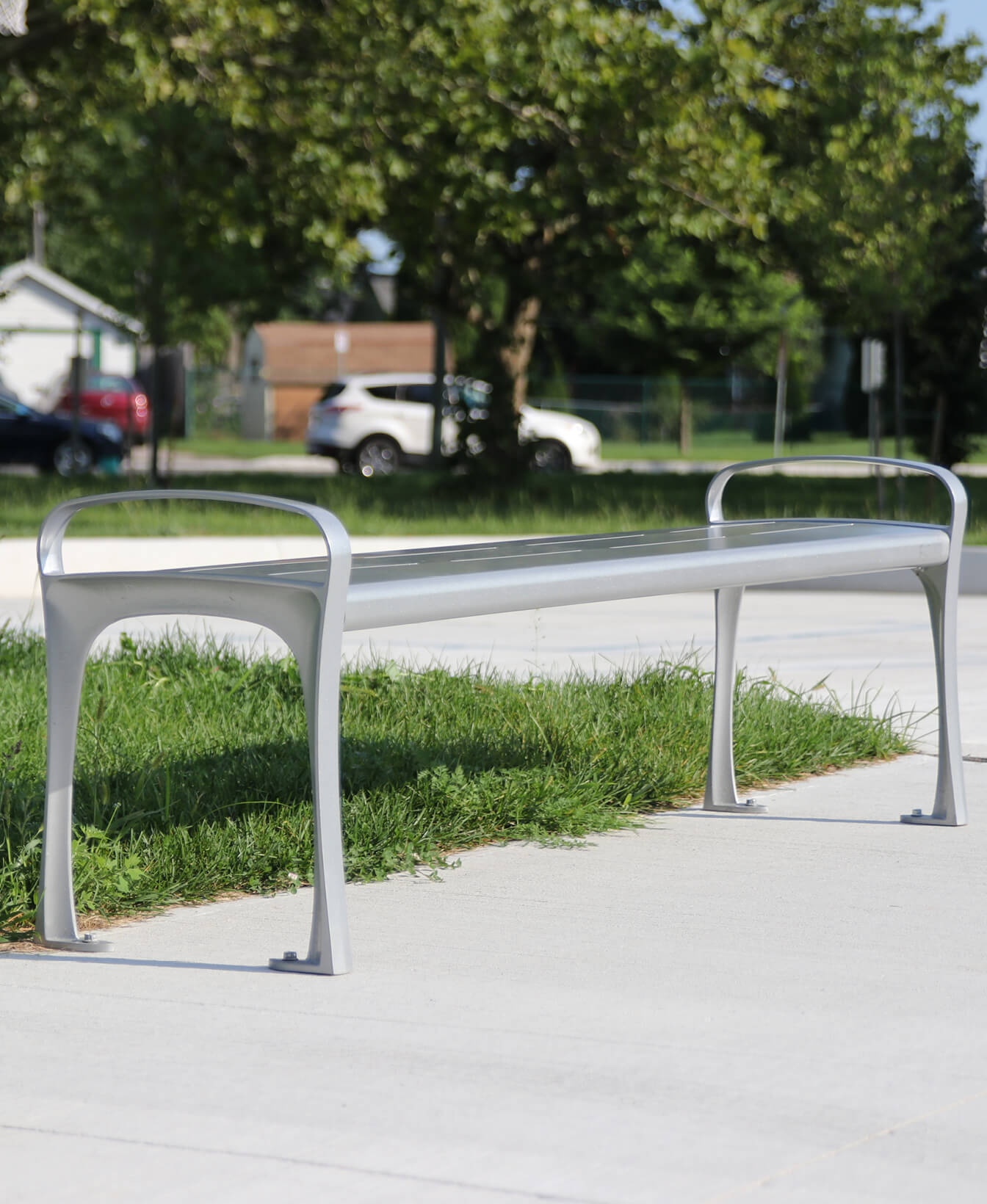 An 870 Backless Bench is placed on a concrete surface next to a grassy area, with trees and parked cars visible in the background.
