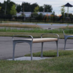 Two empty benches are placed on patches of grass beside a paved path in a park, with trees and a playground visible in the background.
