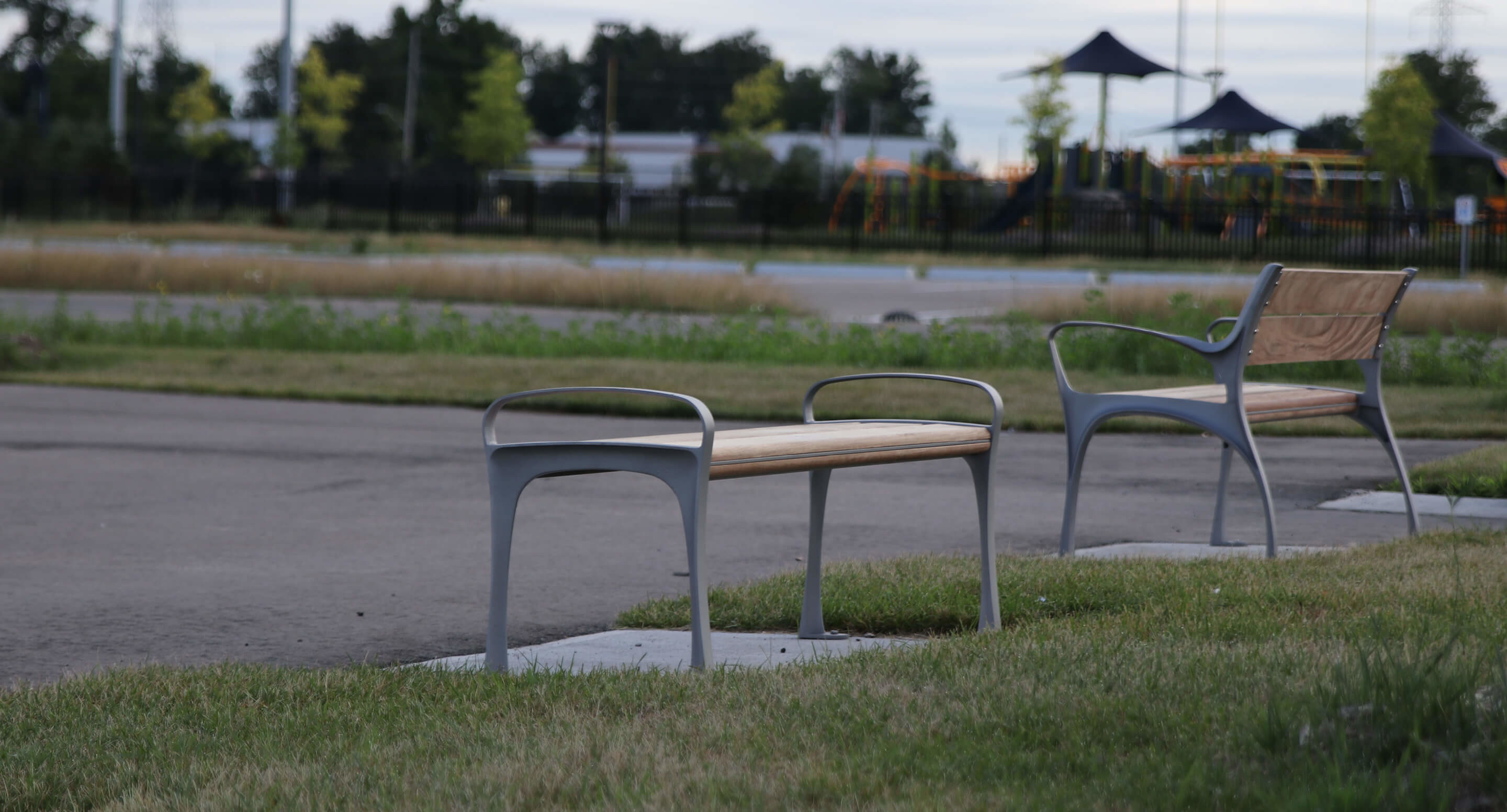 Two empty benches are placed on patches of grass beside a paved path in a park, with trees and a playground visible in the background.