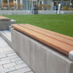 Concrete benches with wooden seats are placed along the edge of a paved walkway beside a grass lawn in an outdoor area near residential buildings.