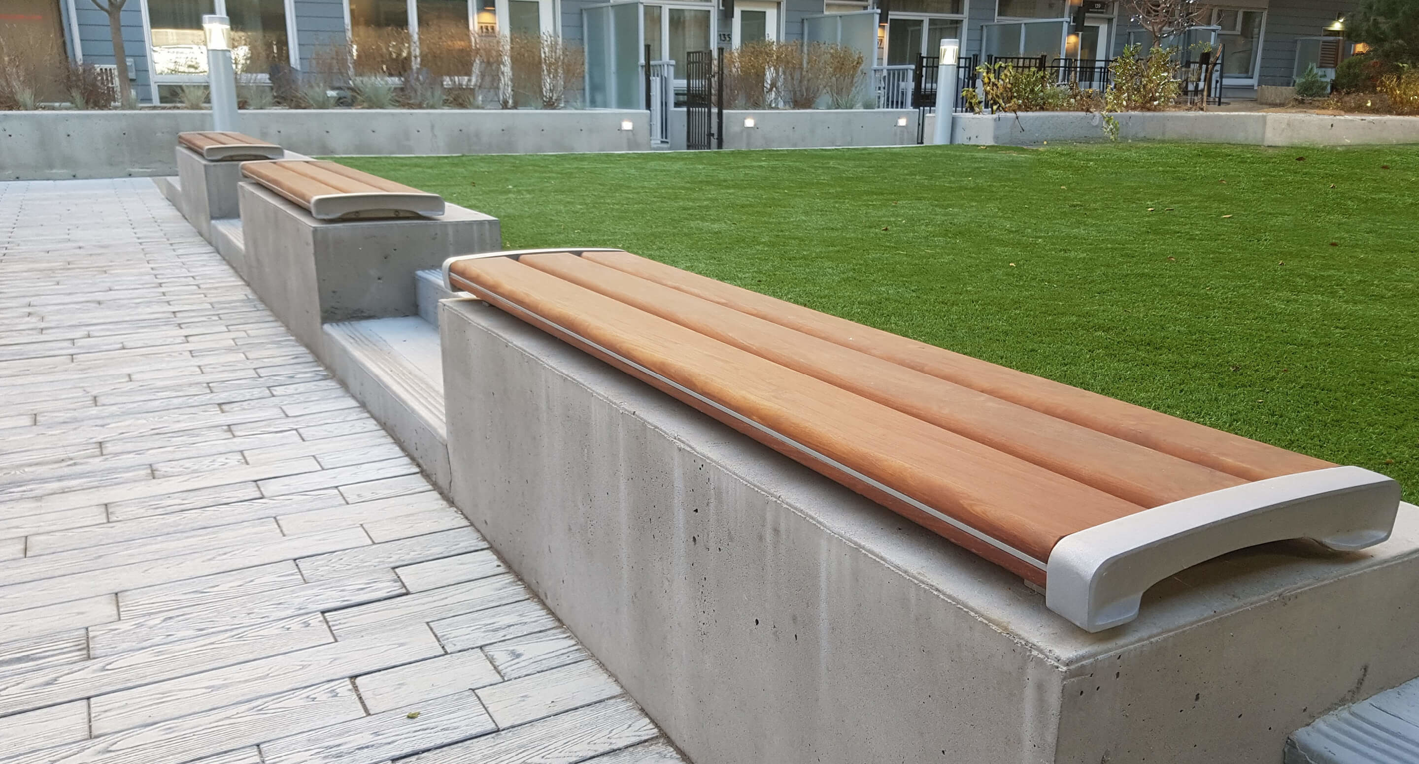 Concrete benches with wooden seats are placed along the edge of a paved walkway beside a grass lawn in an outdoor area near residential buildings.