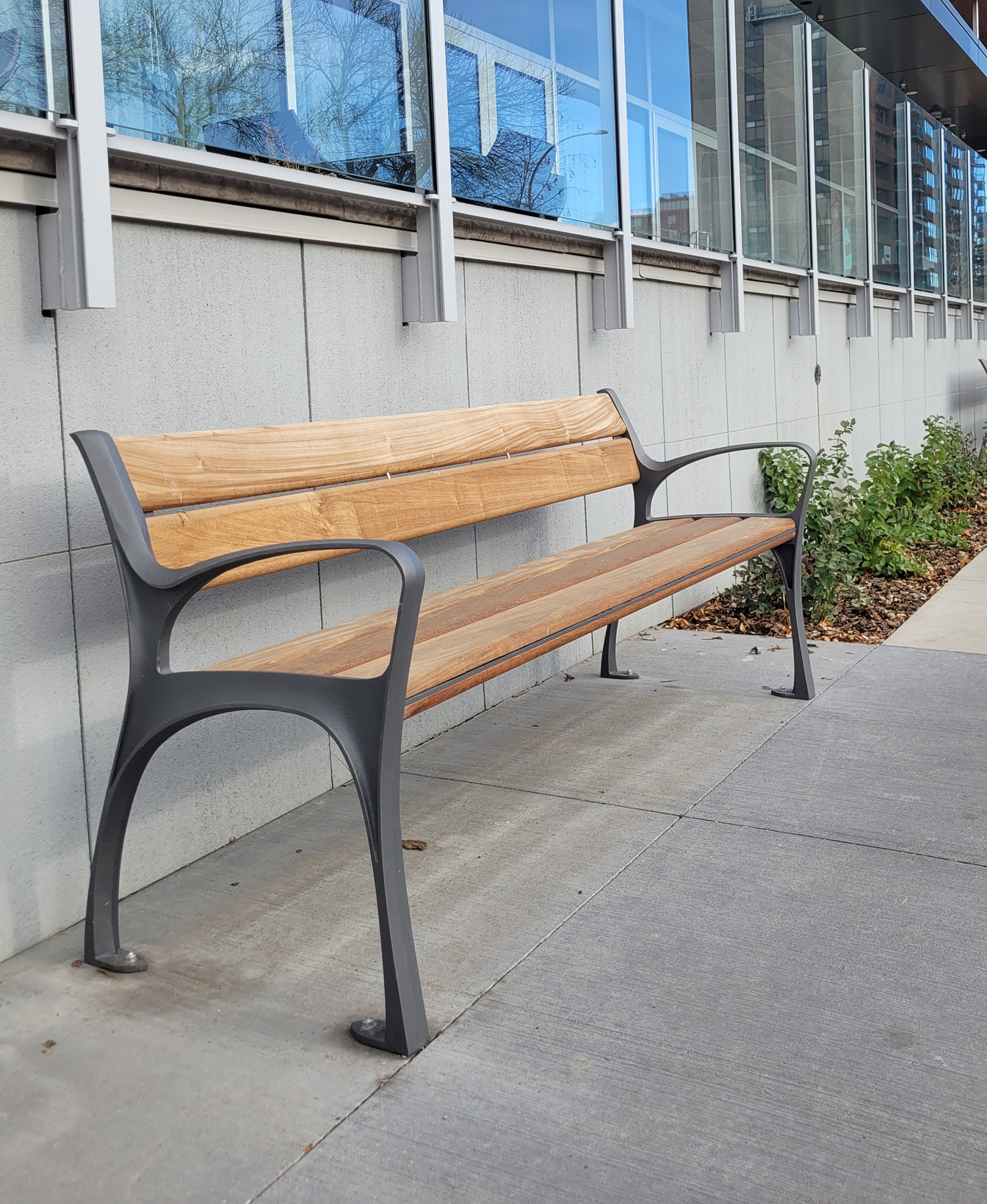 An 870 Backed Bench is placed on a concrete sidewalk beside a modern building with large windows and a landscaped area.