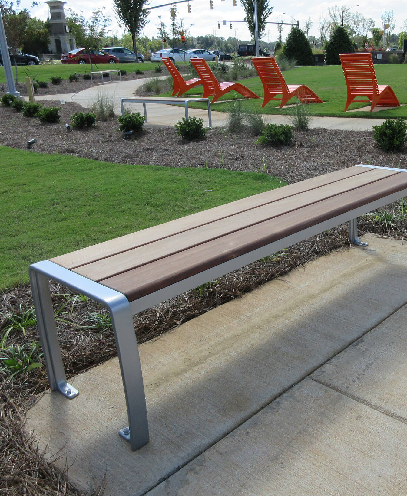 A 970 Backless Bench made of metal and wood sits on a sidewalk near a lawn, with several red lounge chairs and small bushes visible in the background.