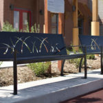 Two metal benches with decorative grass patterns are placed on a concrete sidewalk in front of a building, surrounded by landscaping.