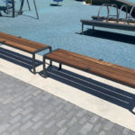 Two wooden benches with metal frames are placed on a concrete strip beside a blue rubber playground surface and gray paving stones.