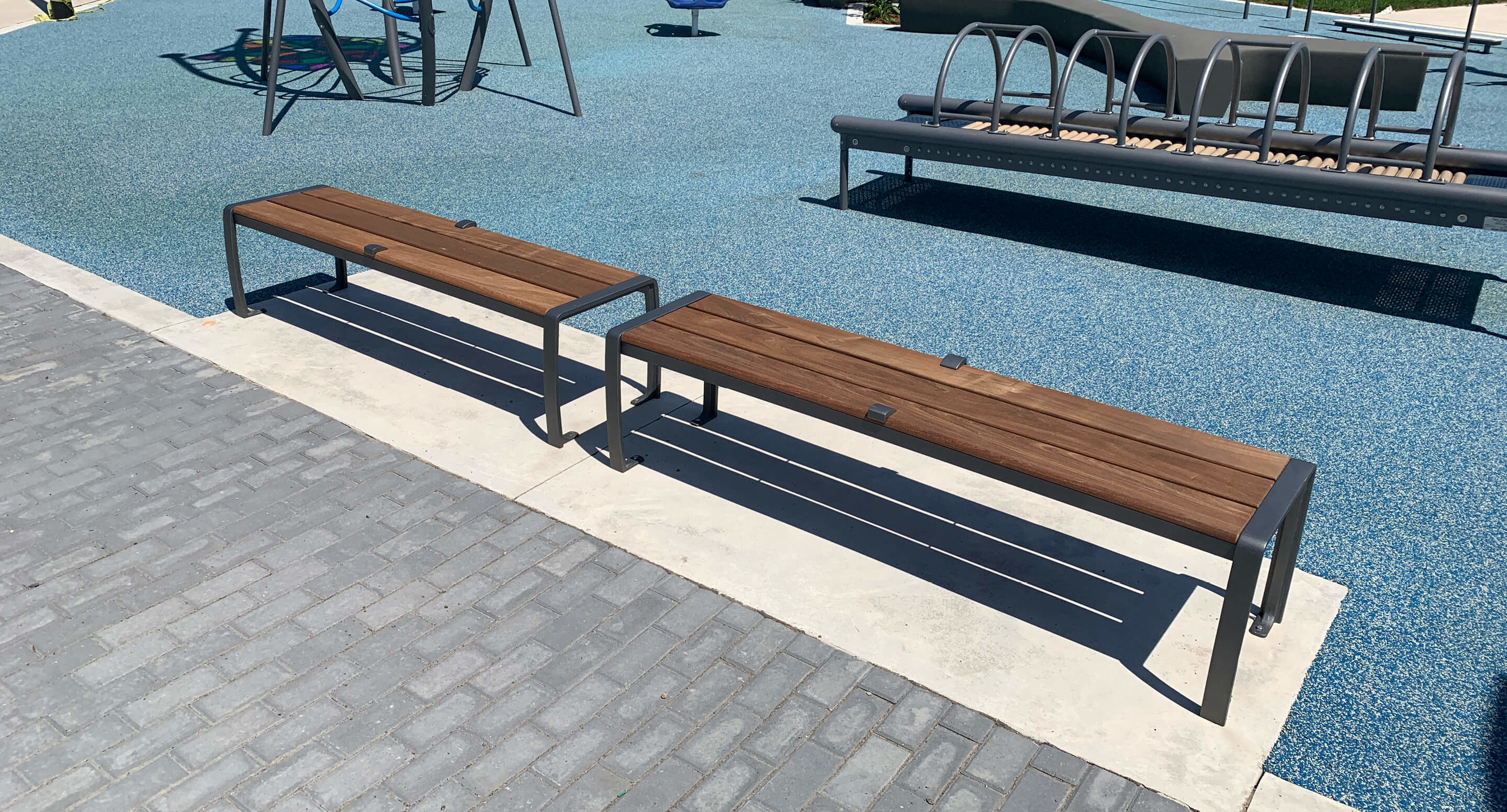 maglin_mbe-0970-backless-bench_1 Two wooden benches with metal frames are placed on a concrete strip beside a blue rubber playground surface and gray paving stones.