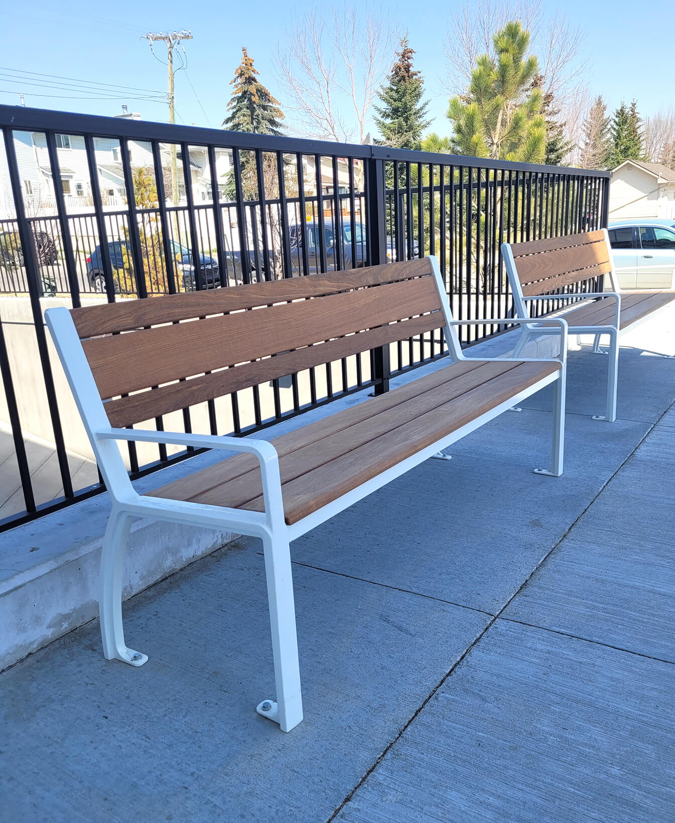 Two 970 Backed Benches with white metal frames are placed on a concrete sidewalk beside a black fence, with trees and parked cars visible in the background.
