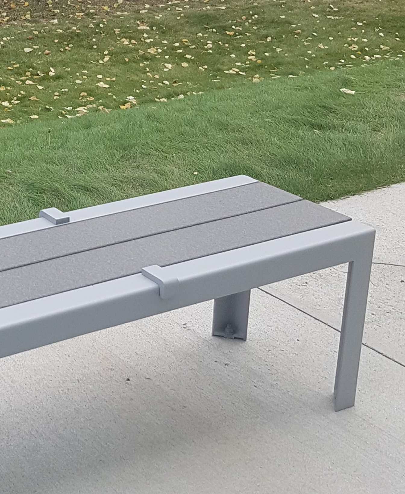 Dispositif anti-planches à roulettes 1050 monté sur un banc en métal gris posé sur du béton, avec de l'herbe verte et des feuilles jaunes dispersées en arrière-plan.