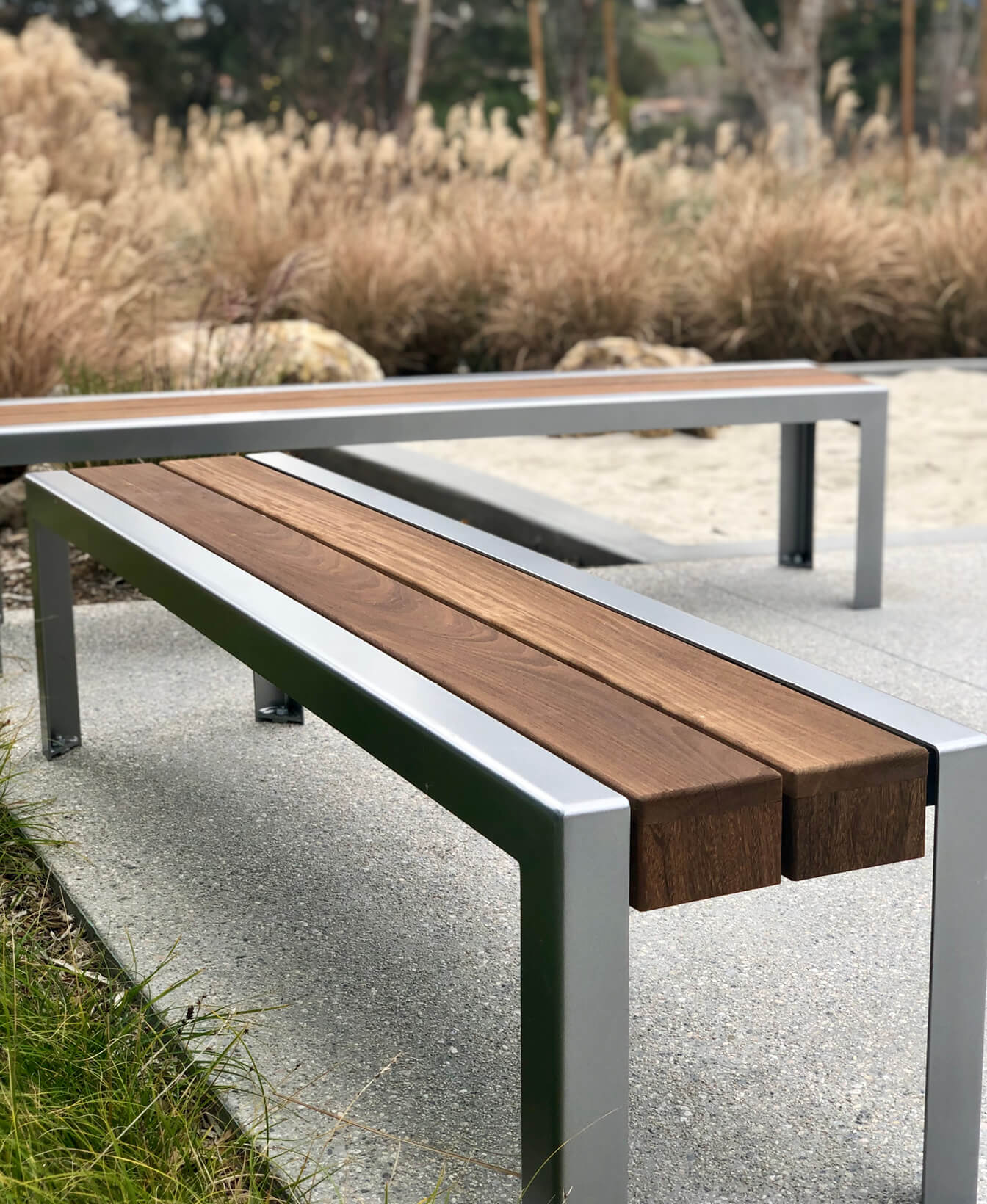 The 1050 Backless Benches, featuring a metal frame and wooden slats, are placed on a concrete surface with grass and ornamental plants in the background.