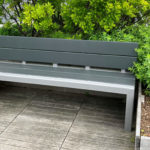 A modern gray bench is positioned on a wooden deck next to green shrubs and plants in an outdoor setting.