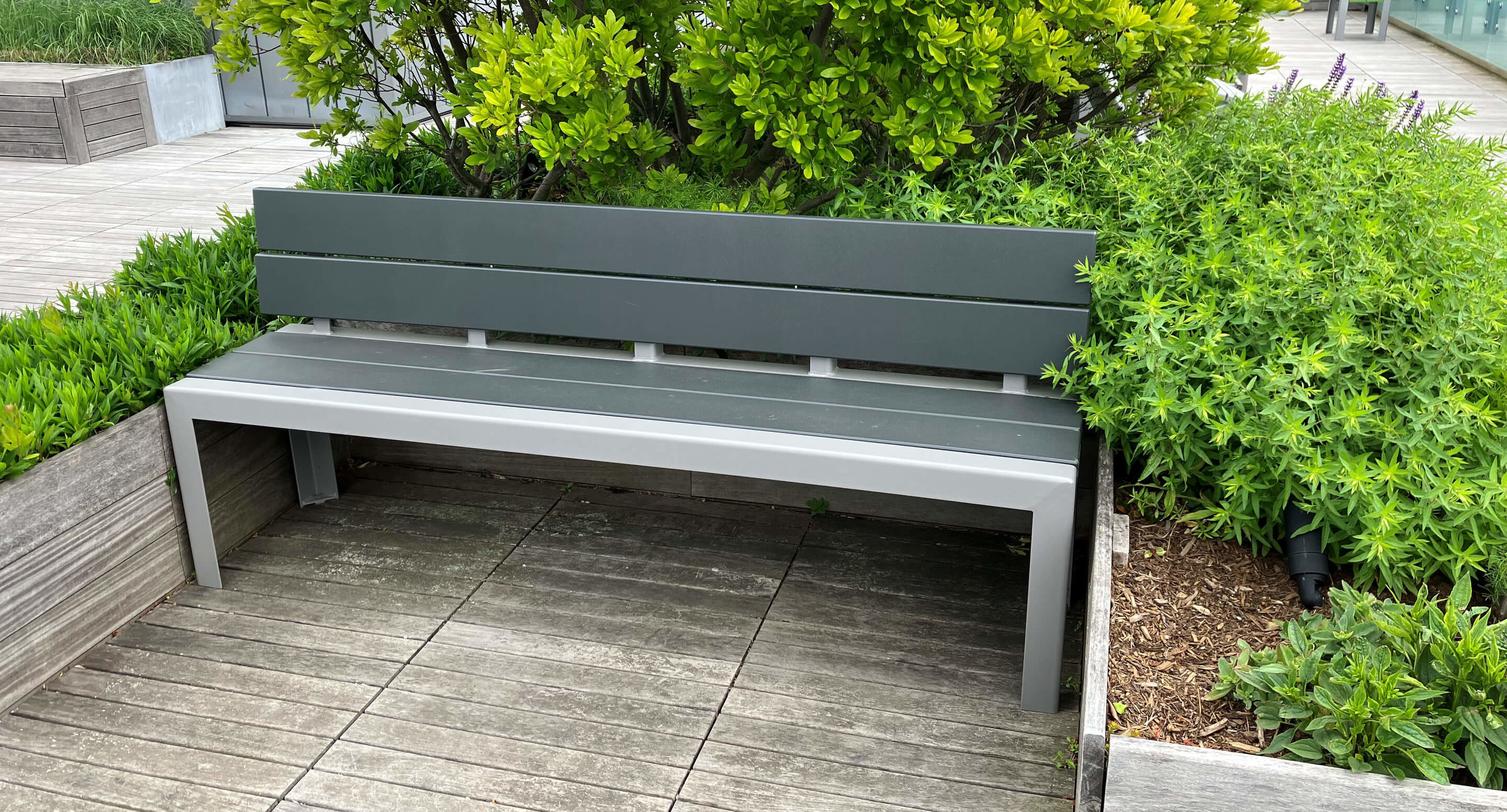 A modern gray bench is positioned on a wooden deck next to green shrubs and plants in an outdoor setting.