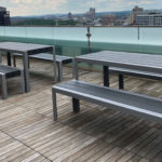 Outdoor rooftop patio with modern gray picnic tables and benches on a wooden deck, overlooking a cityscape with buildings in the background.