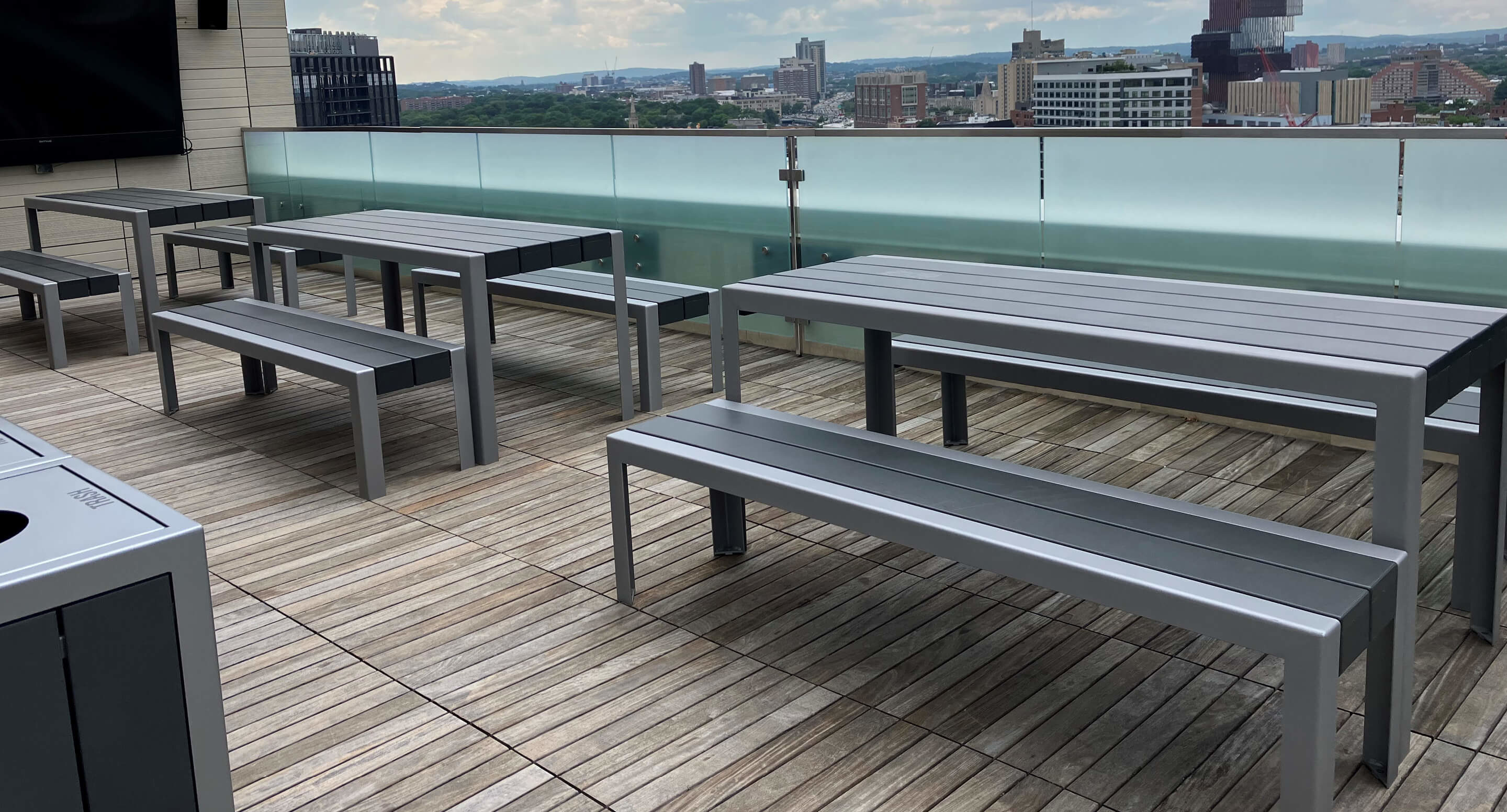 maglin_mbe-1050-table-backless-benches_1 Outdoor rooftop patio with modern gray picnic tables and benches on a wooden deck, overlooking a cityscape with buildings in the background.