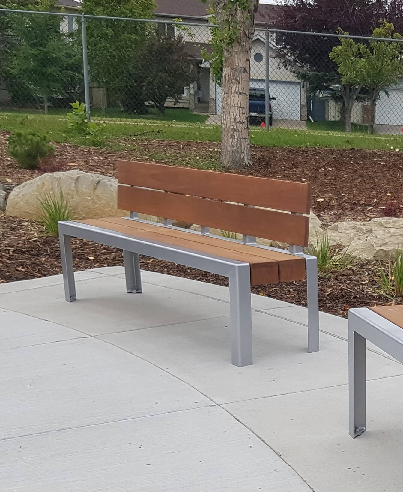 The 1050 Backed Benches, featuring a wooden seat and backrest on a metal frame, are installed on a concrete path in the landscaped park area.
