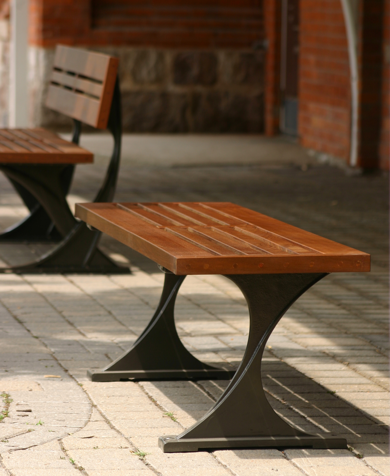 A 1200 Backless Bench made of wood and metal sits on a paved outdoor area, with another matching bench and brick walls in the background.