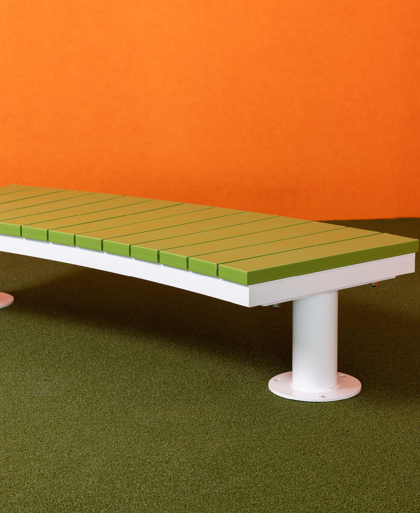 Le Banc Ogden pour enfants, moderne avec des lattes vertes et une base blanche, est posé sur un sol vert devant un fond orange.