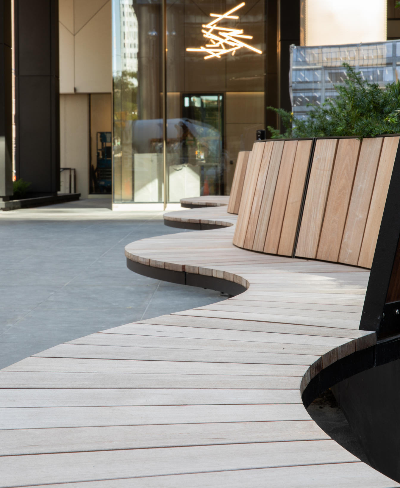 Le Bancs Ogden est un banc en bois courbé au design moderne, présenté sur un espace extérieur pavé à côté d'un bâtiment doté de portes en verre et de luminaires intérieurs géométriques.