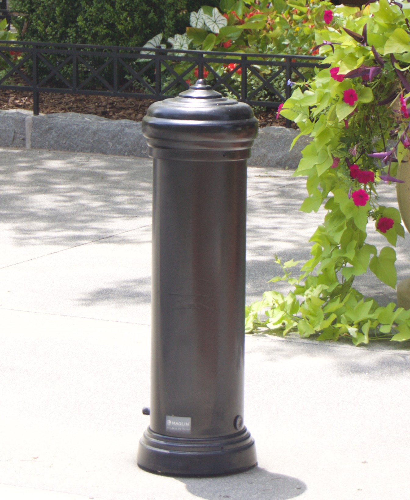 La borne 200, un piédestal cylindrique noir au sommet arrondi, se dresse sur un trottoir en béton près d'un feuillage vert et de fleurs roses.