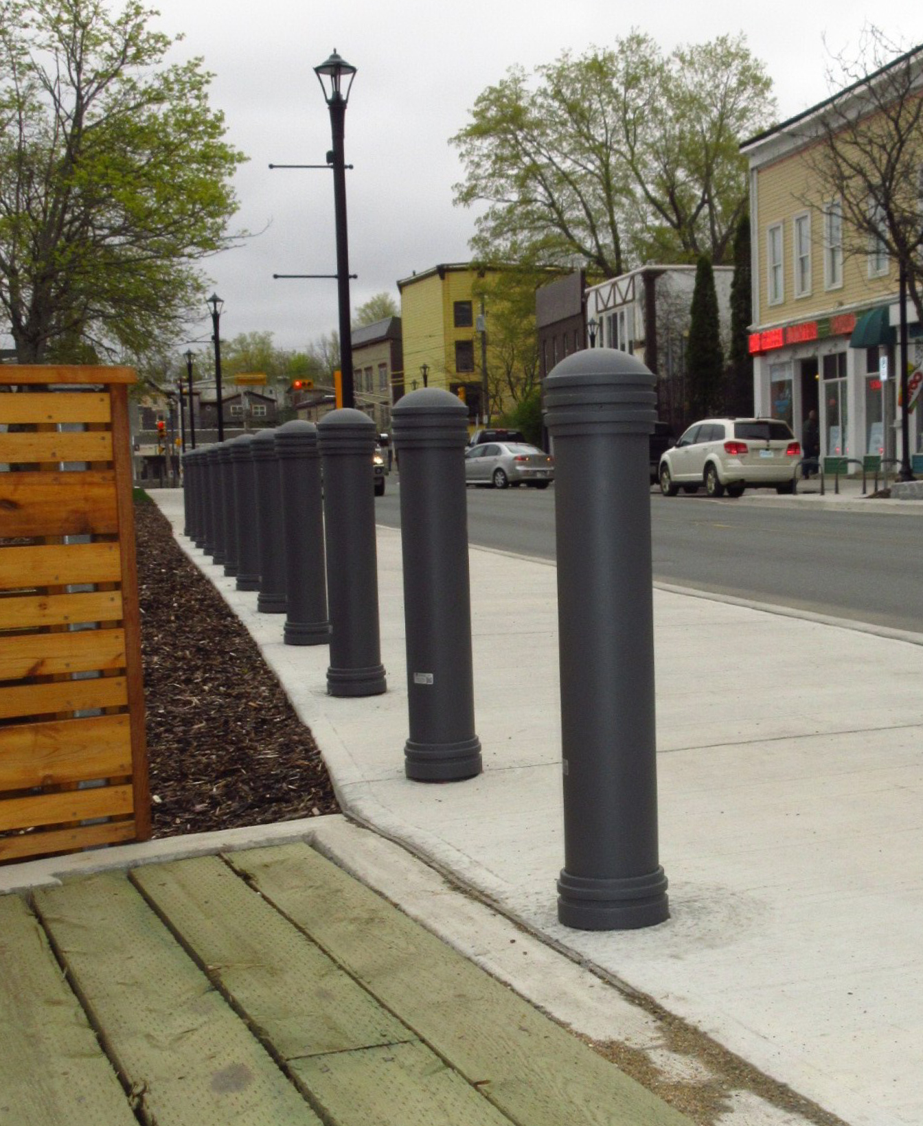 Une rangée de bornes grises Bollard 650 borde le trottoir d'une ville, le long d'une rue, avec des magasins, des voitures en stationnement et des arbres en arrière-plan.