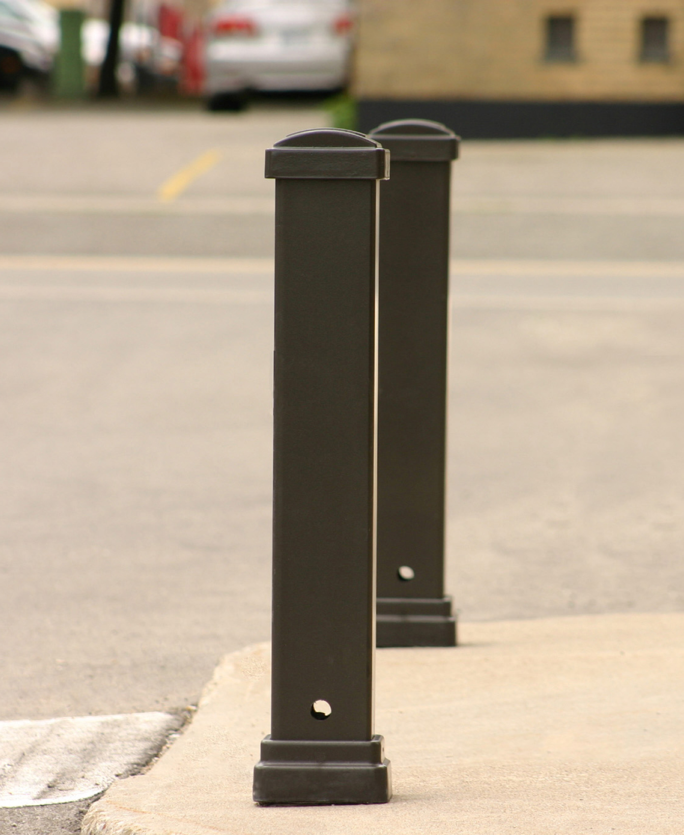 Deux bornes Bollard 970 en métal noir sont installées sur un trottoir en béton près d'une rue, avec des voitures en stationnement et un bâtiment visible à l'arrière-plan.