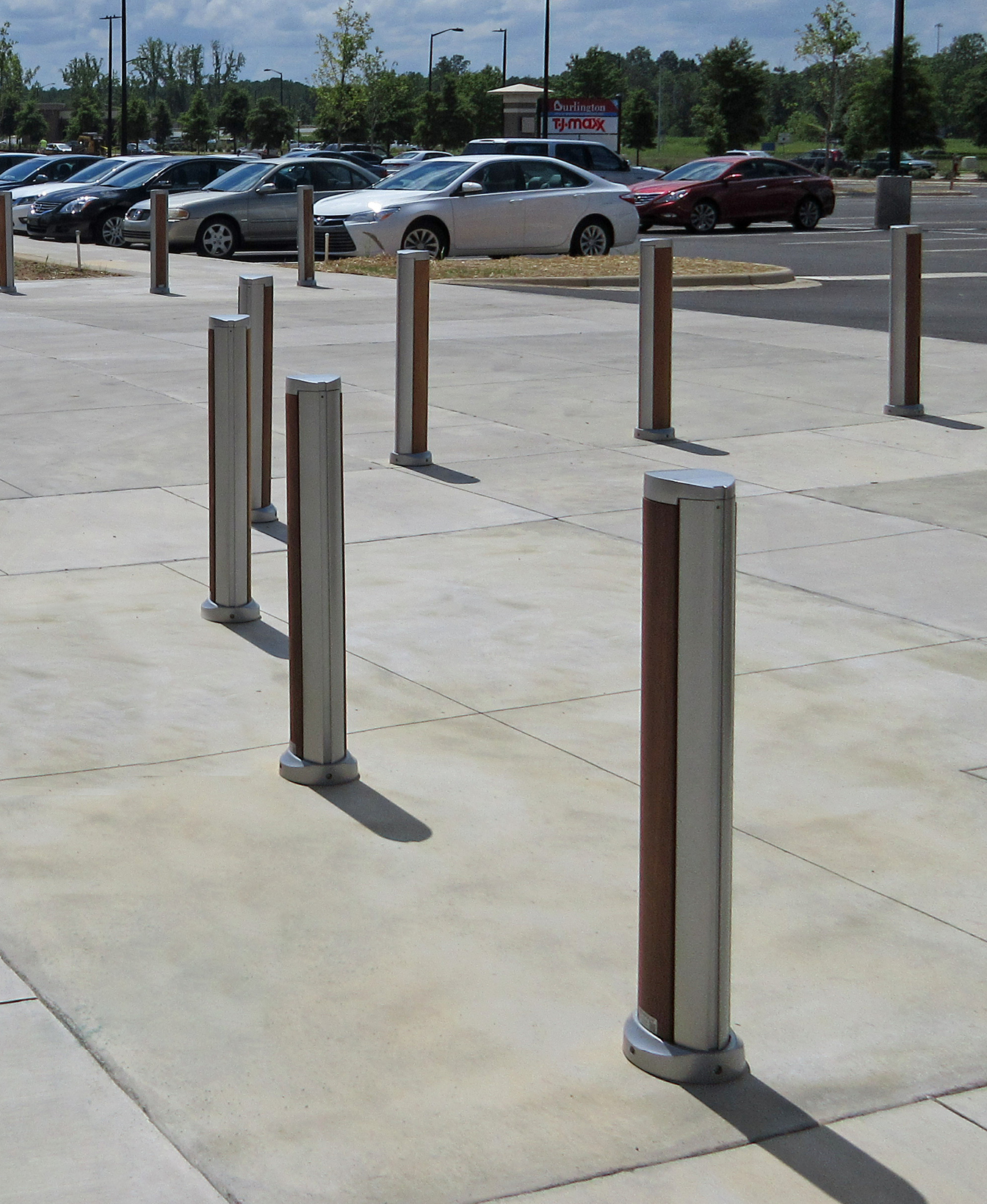 A line of SC Bollards, made of metal and wood, is installed along a paved sidewalk, with a parking lot and several parked cars visible in the background.