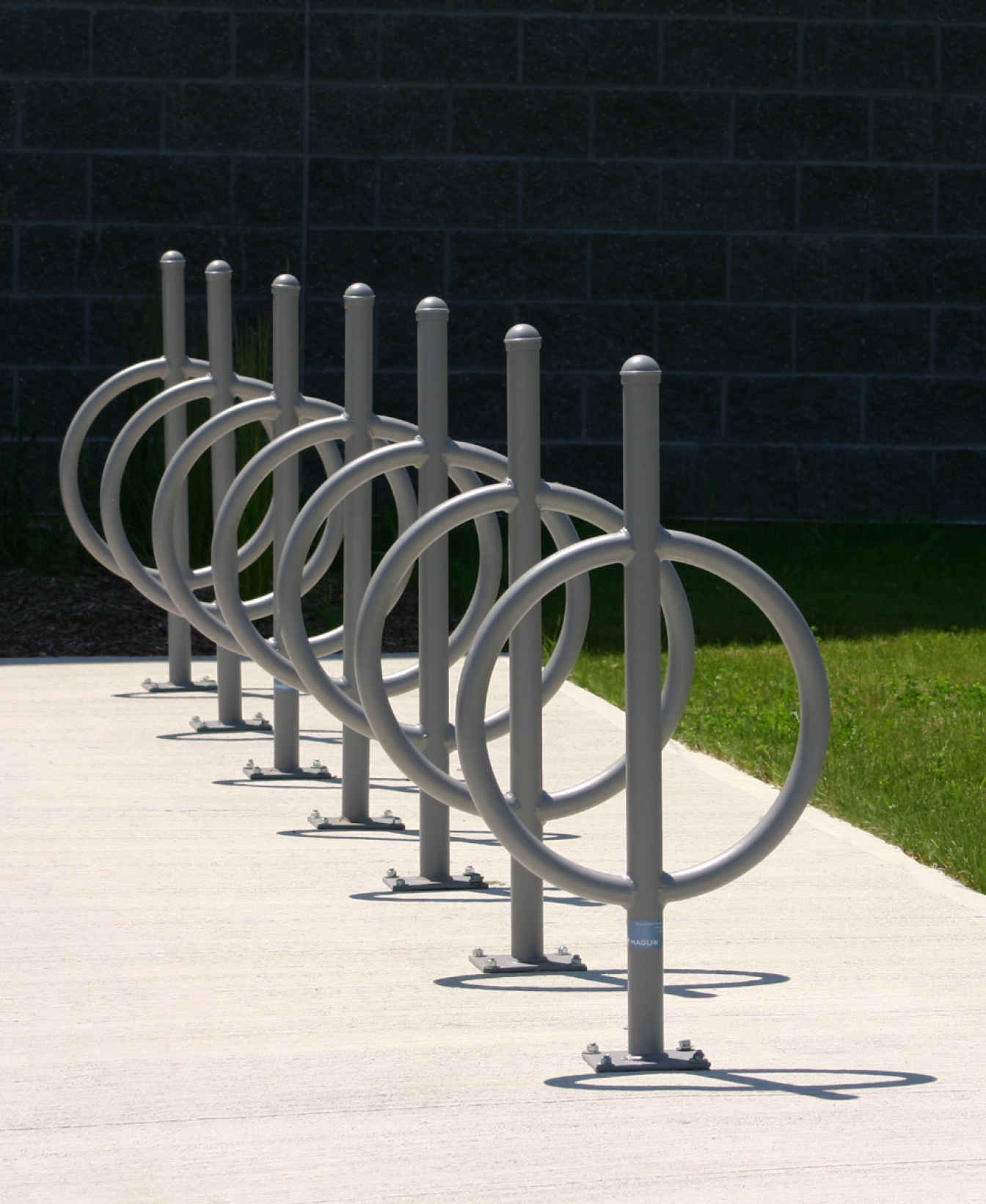 A row of 100 Bike Racks is installed on a concrete sidewalk beside a grassy area and a dark brick wall.