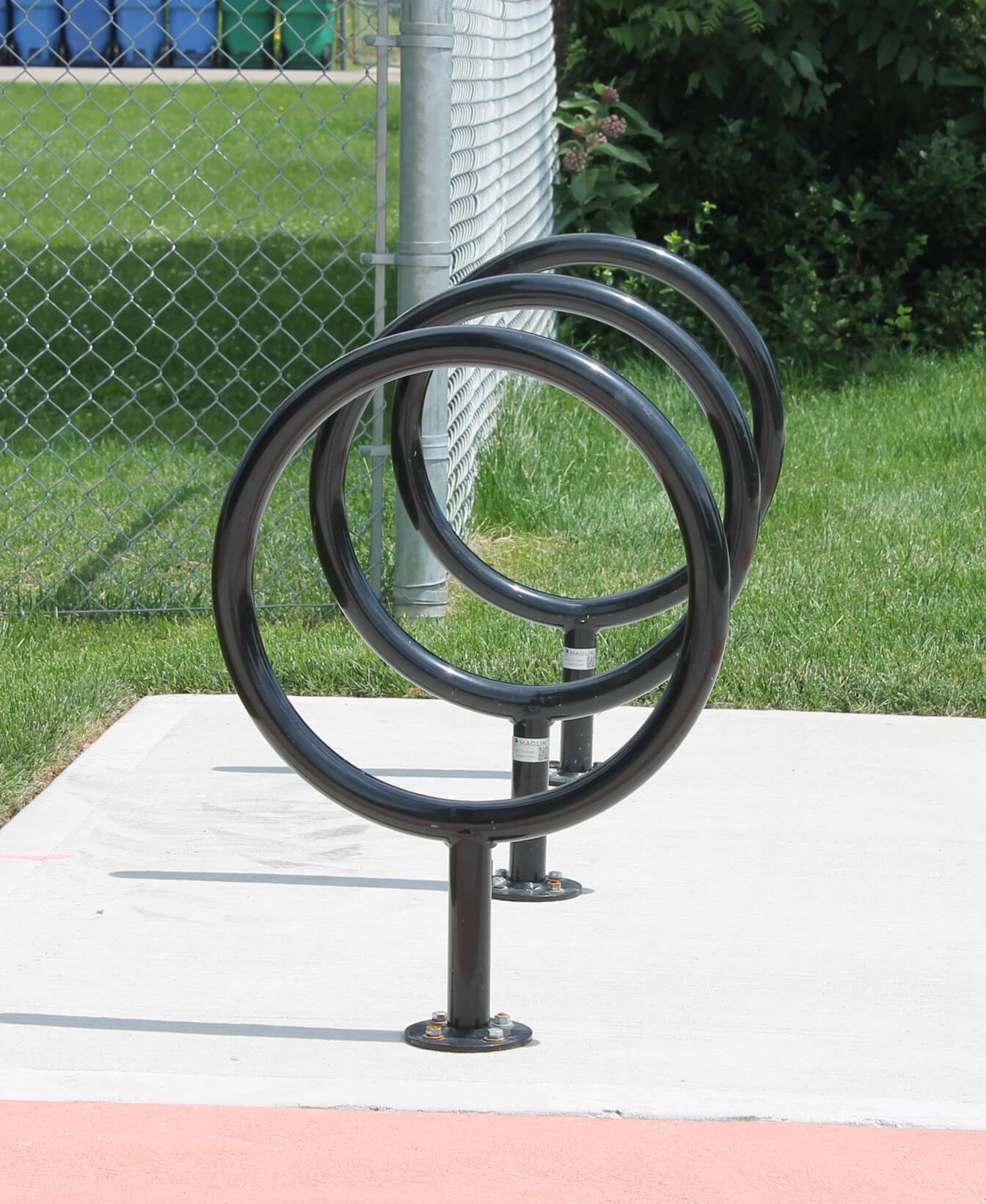 Three 150 Bike Racks, black and circular, are installed on a concrete pad next to a chain link fence and green grass.