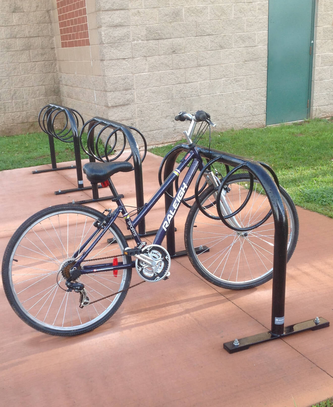 A blue bicycle is parked in a 300 Bike Racks metal rack outside a beige brick building with a green door.
