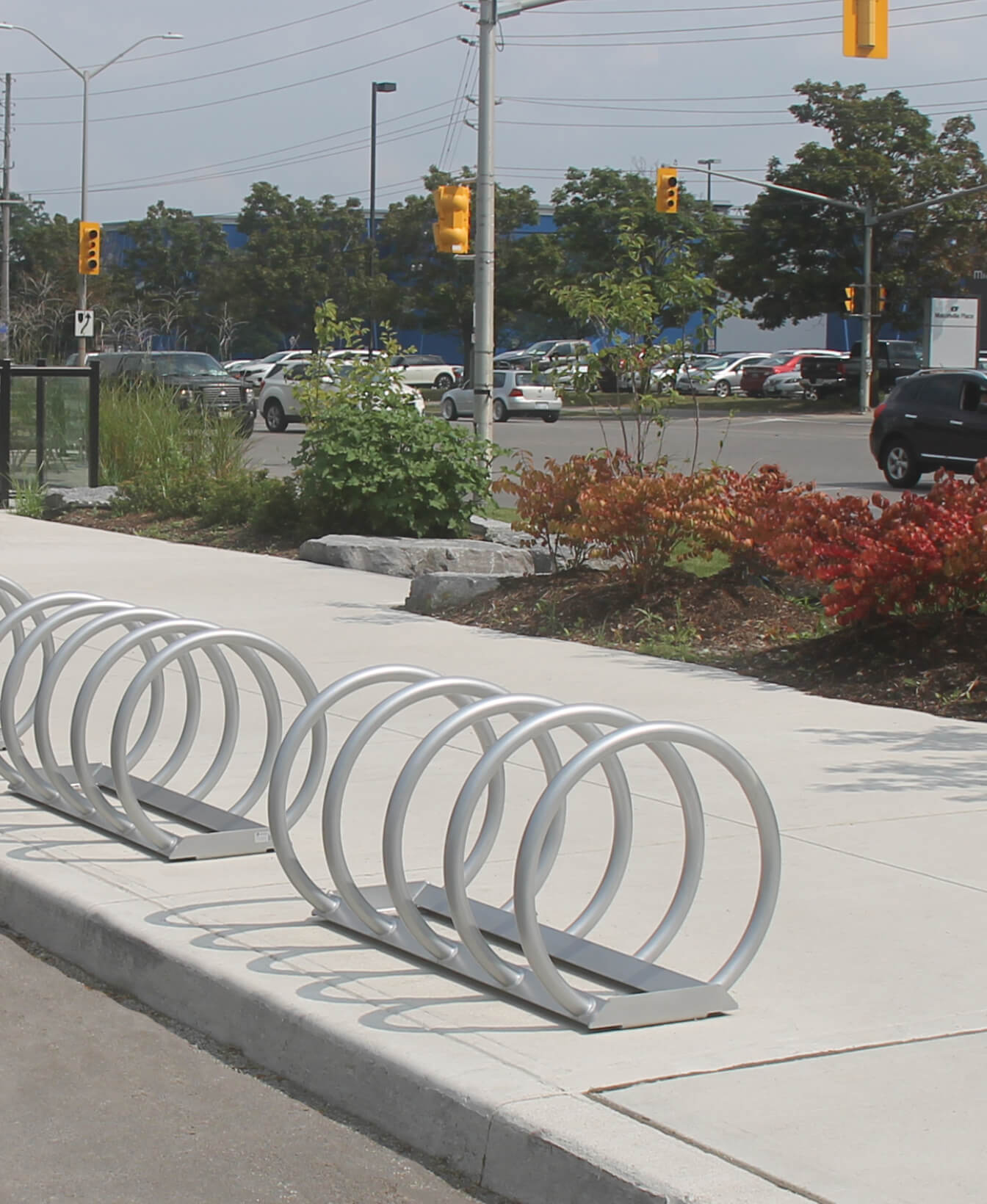 A row of empty 350 Bike Racks is installed on a sidewalk near a parking lot, with traffic lights and trees in the background.