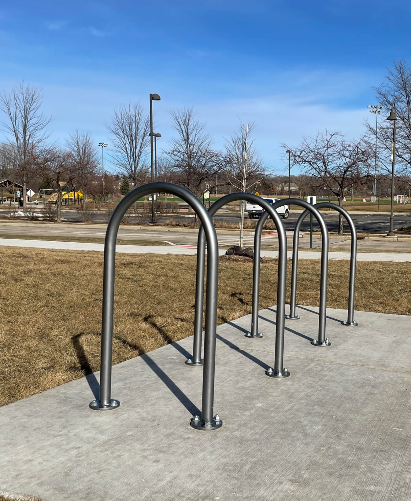 Four 500 Bike Racks are installed on a concrete pad in an outdoor park, with dry grass, bare trees, and a clear sky in the background.