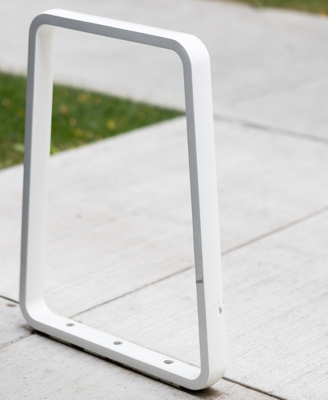 The Iconic Bike Rack, featuring a white metal and rectangular design, is installed on a concrete sidewalk beside a grassy area.