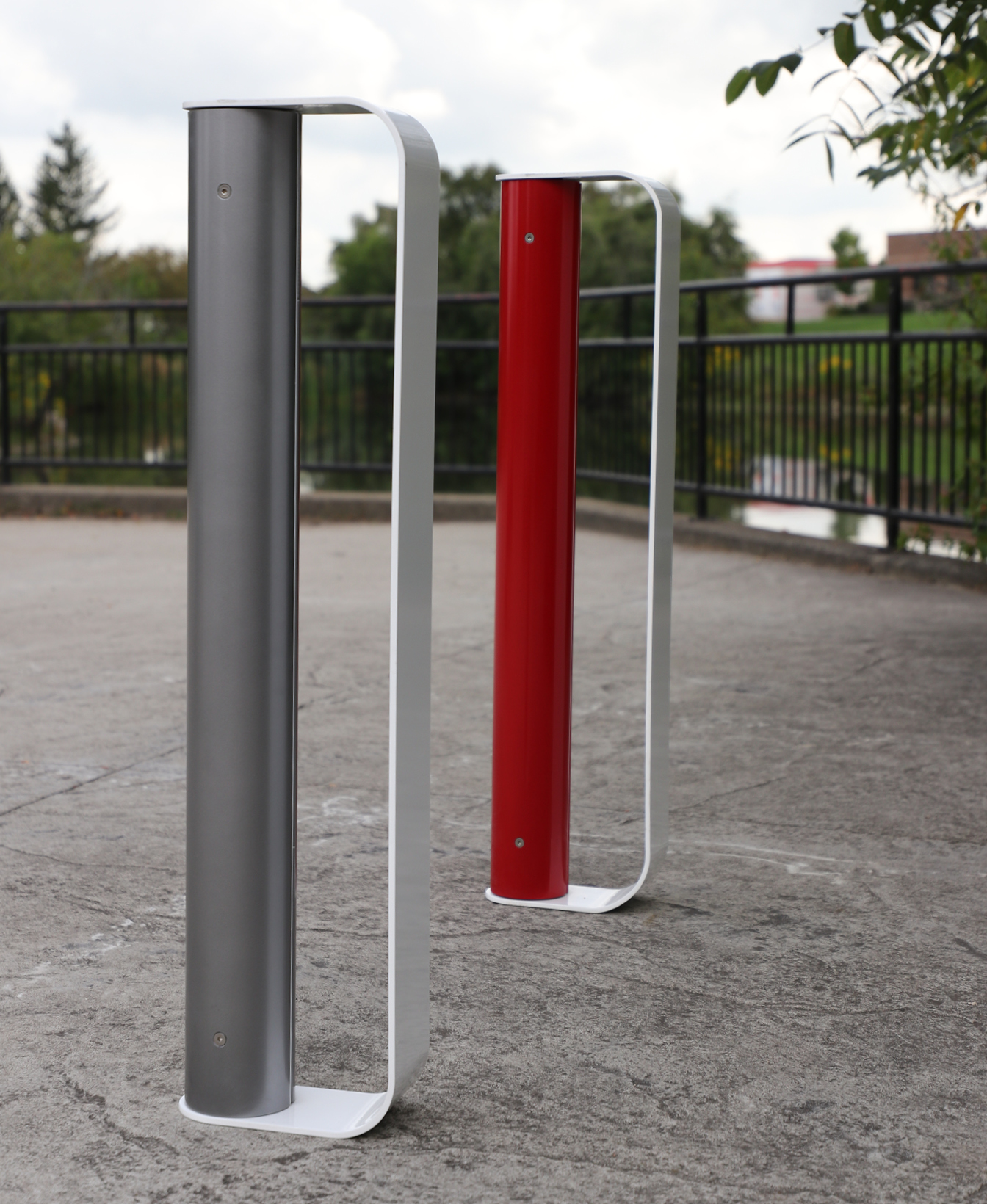 Two MUG Bike Racks, one gray and one red, stand vertically on a paved surface near a black metal fence and trees.